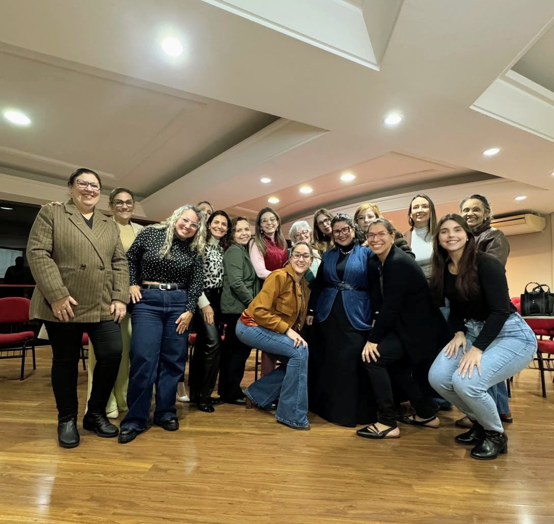 Grupo de mulheres posando em pé e agachadas em salão com piso de madeira e cadeiras vermelhas, na sede da ACIS