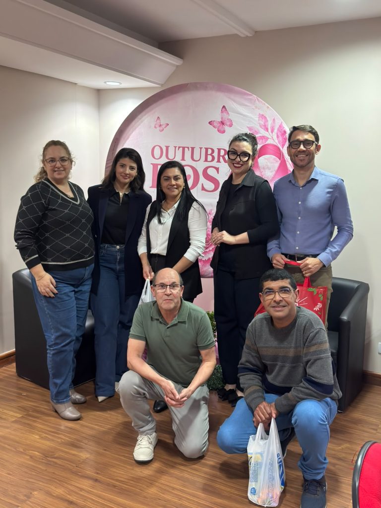 Grupo de pessoas posa em frente a um painel rosa com a inscrição Outubro Rosa e um laço rosa, algumas segurando sacolas.